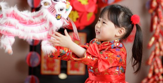 A young girl in traditional Chinese attire smiling and playing with a festive lion dance puppet during Lunar New Year celebrations.