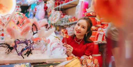 Young woman in traditional attire exploring a festive market decorated with colorful ornaments and Lunar New Year decorations.