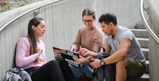 A diverse group of students sitting on outdoor stairs, practicing English vocabulary and exchanging new words using a laptop.