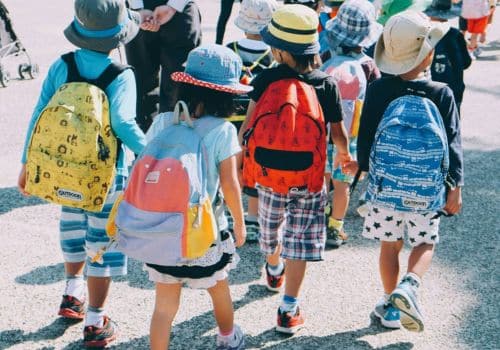 Young children on an educational tour with colorful backpacks, led by a teacher.