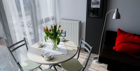 A bright and modern apartment dining area with a round table, chairs, fresh flowers, and a cozy seating corner with red cushions.