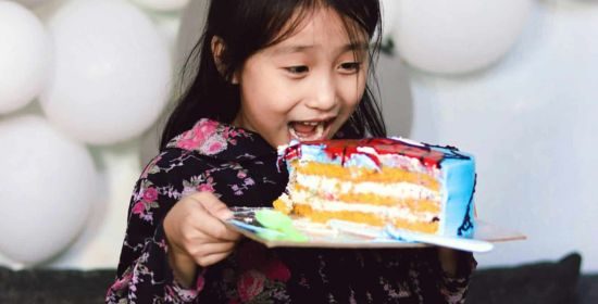 A young girl excitedly holding a colorful slice of cake during a celebration, surrounded by white balloons.